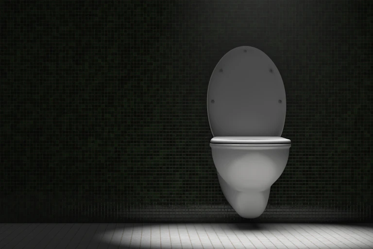 A spotlight shines from above on a white toilet, mounted on a dark wall