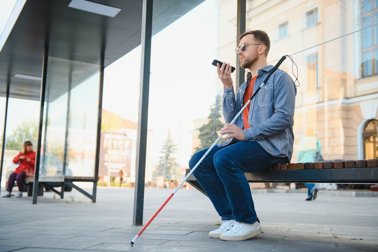 a man sitting at a bus stop holding a cane and speaking into a mobile phone