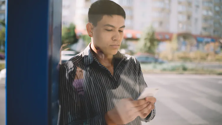 a young man in a leather jacket looks at his phone while waiting at the bus