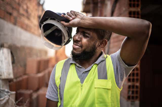 A worker holding his hardhat wipes his forehead with the back of his arm. 