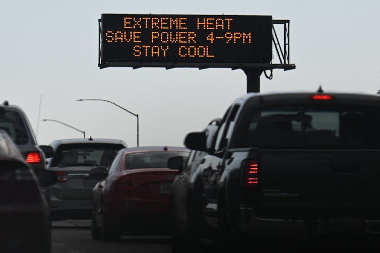 Cars drive under a sign reading: Extreme heat. Save Power 4-9PM. Stay Cool