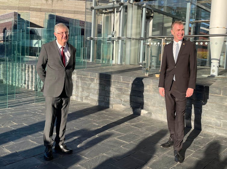 Two men in suits stand smiling in sunlight against a slate floor and glass building in the background.