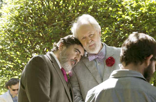 Alfred Molina and John Lithgow resting heads on each other while wearing suits. 