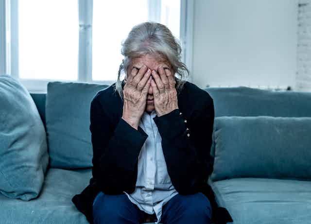 A senior woman sits on a couch with her face in her hands.
