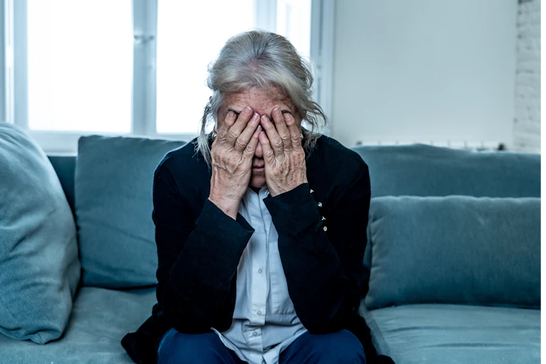 A senior woman sits on a couch with her face in her hands.