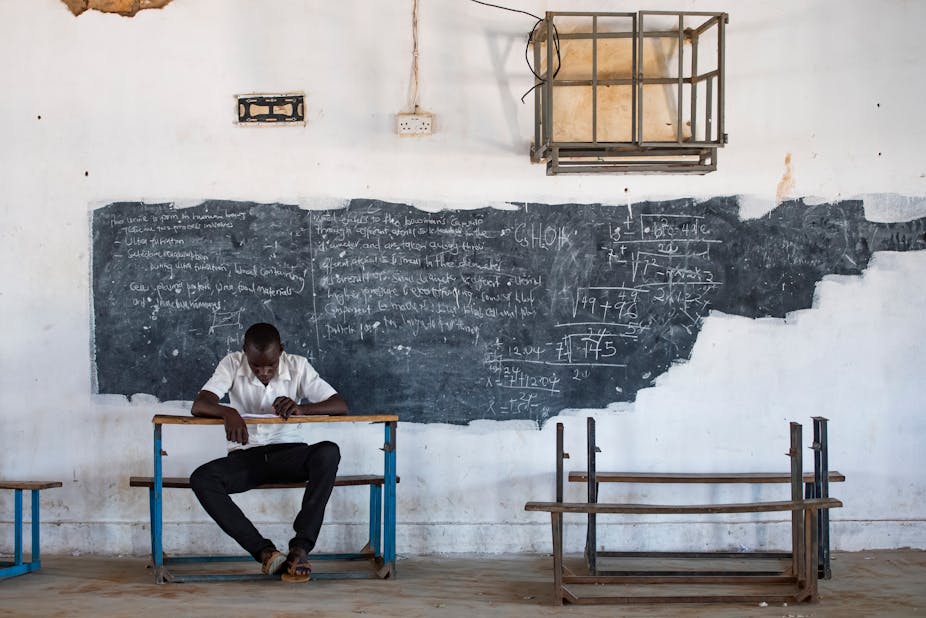A man sits at a school desk in front of a chalkboard covered in writing