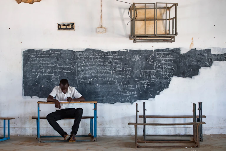 A man sits at a school desk in front of a chalkboard covered in writing