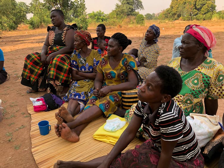 A group of 7 women, two children and a man sit on a mat, talking with someone out of the frame