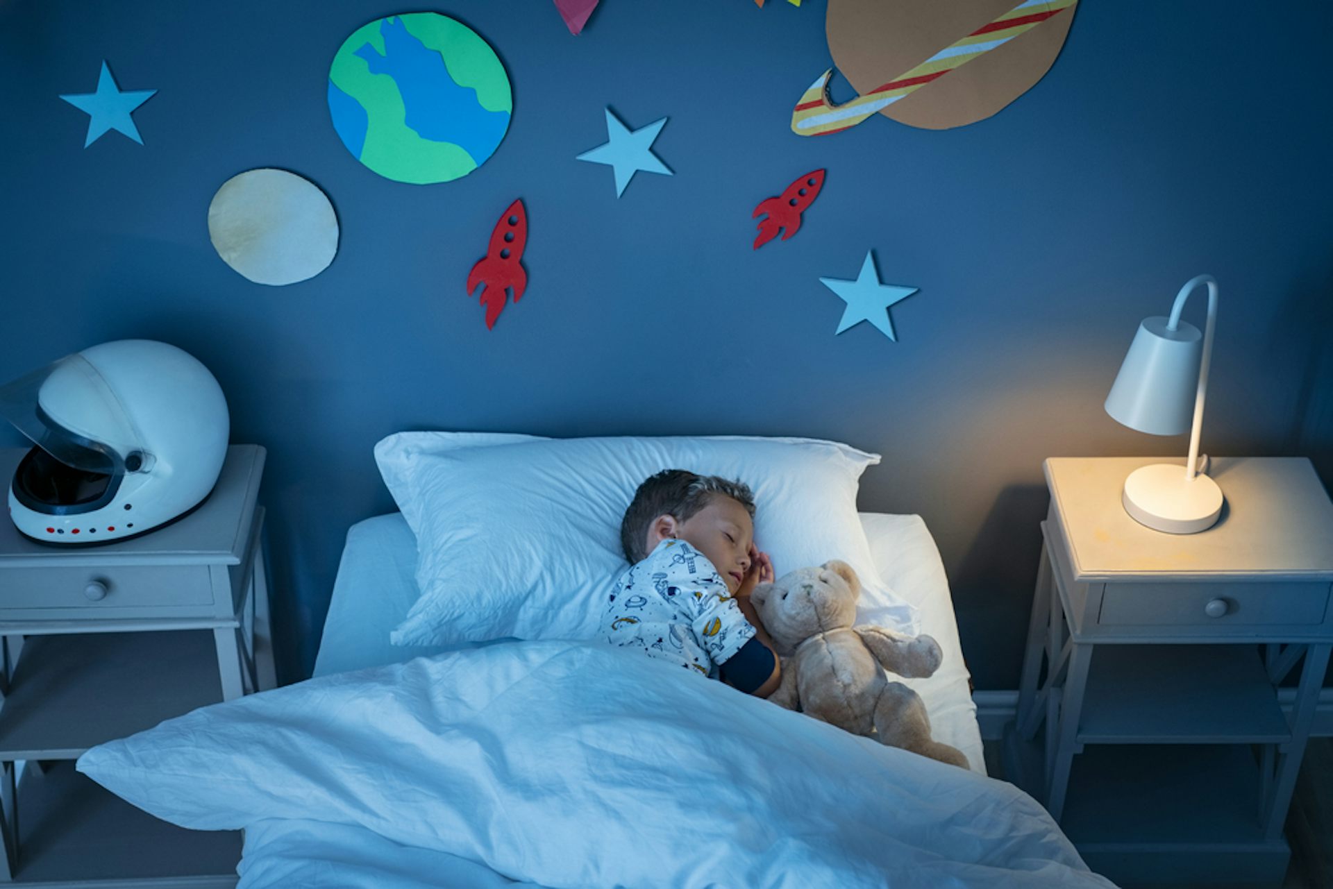 Petit garçon rêvant de devenir astronaute en dormant avec un ours en peluche dans une chambre décorée avec des stickers