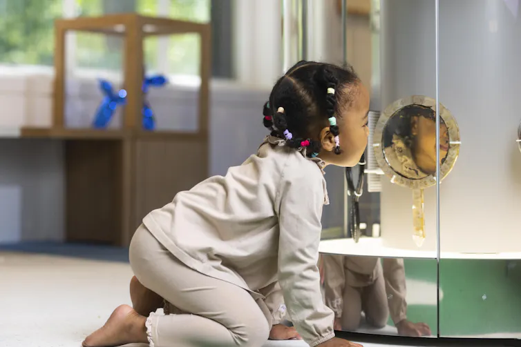 A young black girl with her hair in colourful hairbands bending down to look in a mirror in a display case.