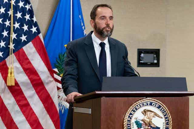 A dark haired, bearded man in a dark suit, white shirt and tie stands at a lectern in front of the US flag and speaks.