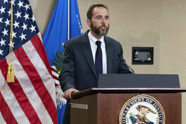 A dark haired, bearded man in a dark suit, white shirt and tie stands at a lectern in front of the US flag and speaks.