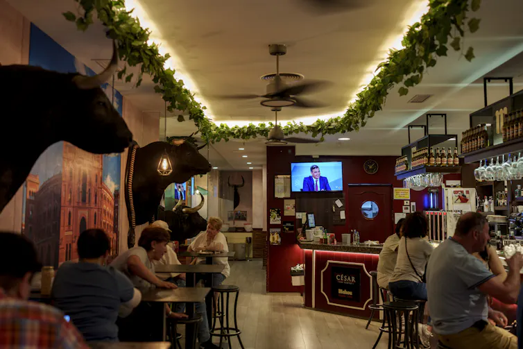 Scène dans un bar, avec des gens attablés et un écran de télévision allumé