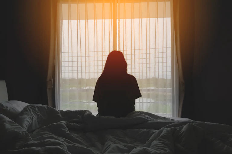 A slihouette of a person sitting on a bed looking towards a window covered in net curtains.