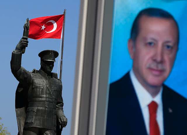 A portrait photo of Recep Tayyip Erdogan  is seen next to a flag of Turkey and a bronze statue of a soldier or man wearing a hat.  