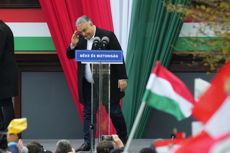 Viktor Orban salutes to a crowd of people while he stands on a podium, with red, white and green flags around him.