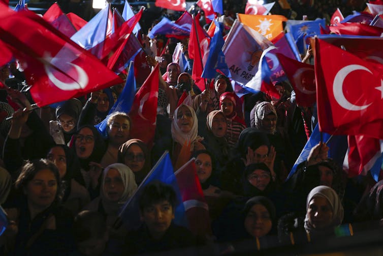 A group of people, some of whom are wearing headscarves, wave Turkish flags and appear to celebrate.