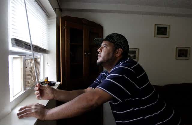 A man opens the shade on an older apartment window with a window air conditioner in it.