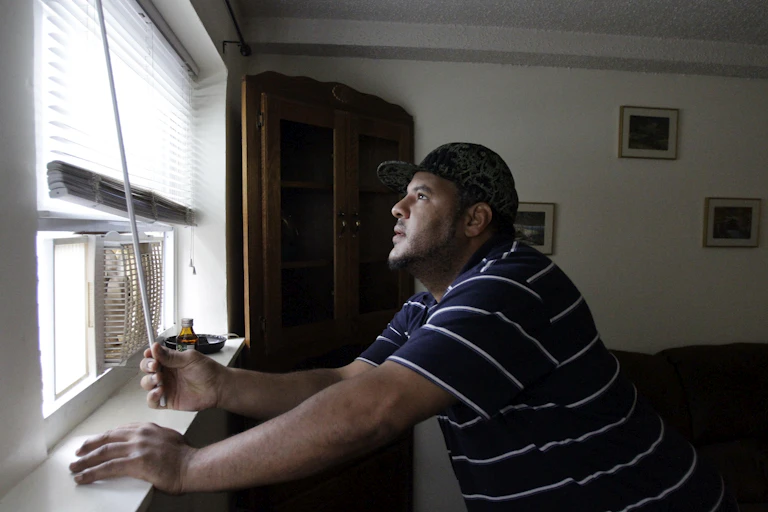 A man opens the shade on an older apartment window with a window air conditioner in it.