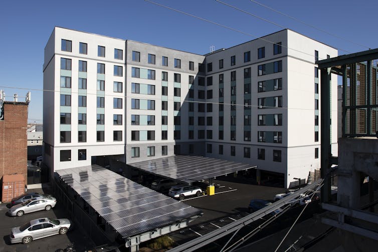 A large apartment building with solar panels on the roof and on shades over the parking area.