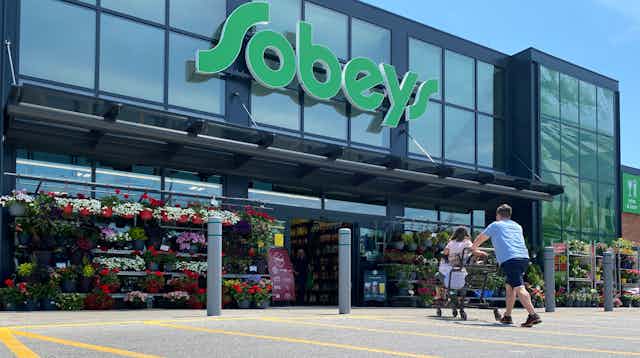 A man pushes a grocery cart with two children sitting in it towards the entrance of a Sobeys grocery store