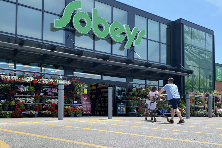 A man pushes a grocery cart with two children sitting in it towards the entrance of a Sobeys grocery store