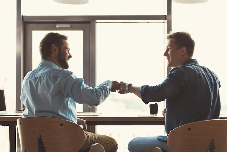 Two men in business casual attire bump fists while chatting over coffee