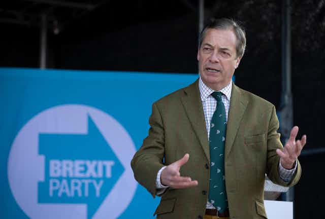 Nigel Farage giving a speech in front of a Brexit Party sign.