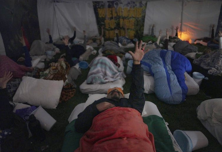 Around a dozen people lie in sleeping bags in a tent, each raising one hand toward the roof.