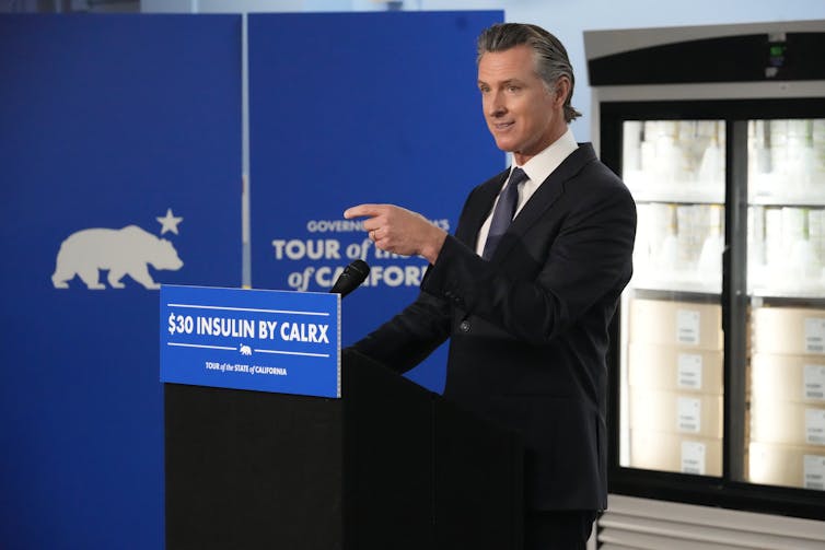 A man in a suit points in front of a lectern that says $30 insulin, with fridges of insulin in the background.