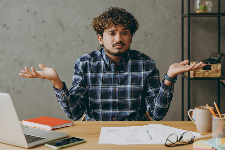 A young man shrugging and looking confused while sitting at a desk in a casual office space