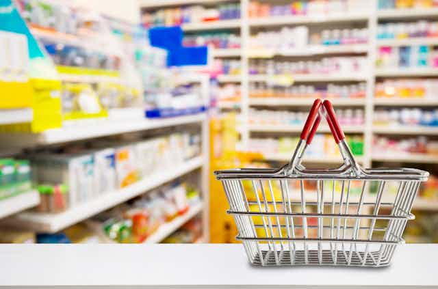 An empty shopping basket sits on the counter at a pharmacy with blurred shelves of medicine and vitamin supplements in the background.