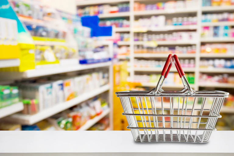 An empty shopping basket sits on the counter at a pharmacy with blurred shelves of medicine and vitamin supplements in the background.