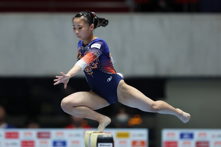 Woman gymnast balancing on one bent leg on a balance beam with the other leg extended straight behind her.