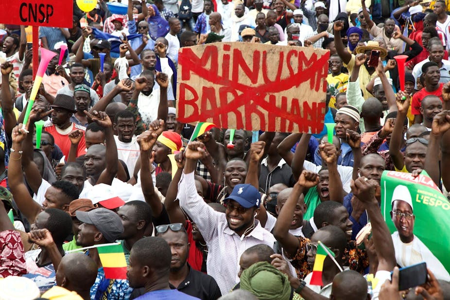A crowd of people holding up their hands in protest, waving green, yellow and red flags, with a poster held up in the middle with the words Minusma and Barkhane written in red and then crossed out