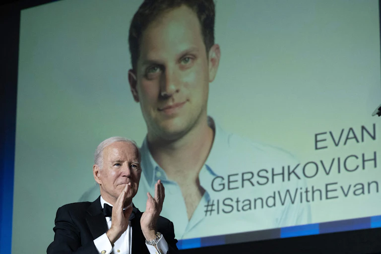As an image of a man with brown wavy hair with the words, ‘I stand with Evan,’ hangs overhead, a gray-haired man in a black suit jacket and bowtie claps his hands.