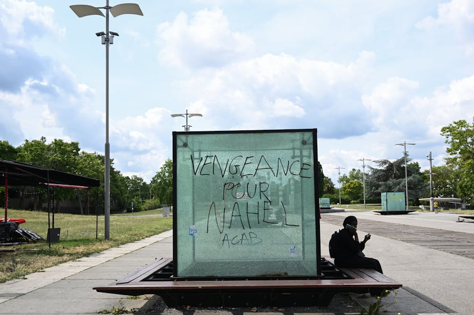Une personne consulte son smartphone à côté d'une vitre portant l'inscription « Vengeance pour Nahel. ACAB »