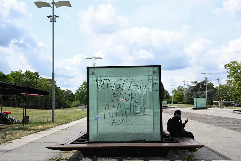 Une personne consulte son smartphone à côté d'une vitre portant l'inscription « Vengeance pour Nahel. ACAB »