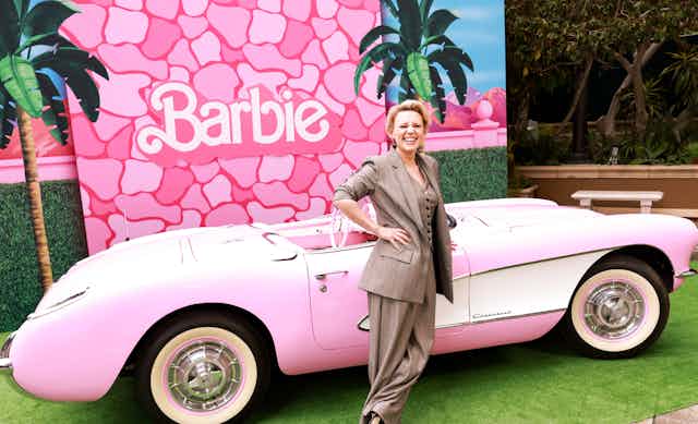 A woman in a beige pantsuit leans against a pink car with a Barbie backdrop.