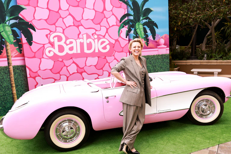 A woman in a beige pantsuit leans against a pink car with a Barbie backdrop.