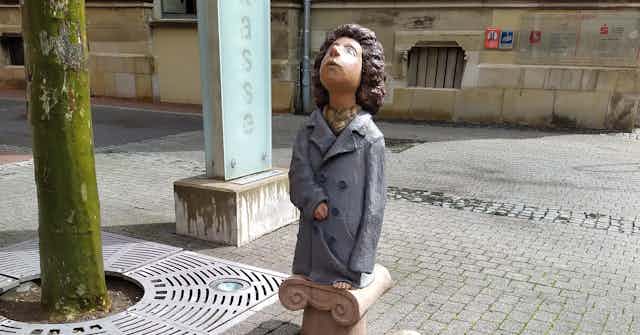 Escultura de una niña vestida con un abrigo que le llega a los pies y que mira al cielo.