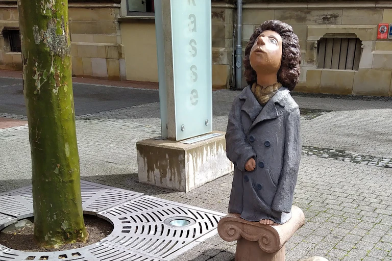 Escultura de una niña vestida con un abrigo que le llega a los pies y que mira al cielo.