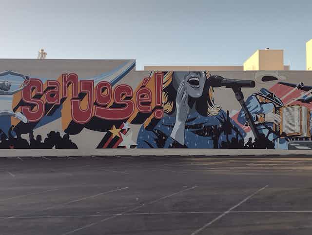 A mural reads "san jose!" and shows a woman singing into a microphone. In the foreground is a large empty parking lot.