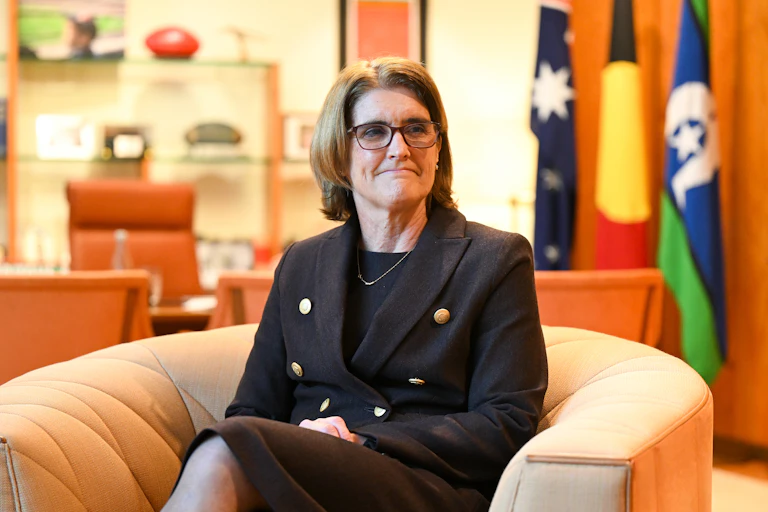 woman in glasses and suit sits with flags behind her