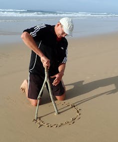 A man holding a forked stick kneels in the sand on a beach, drawing a circle