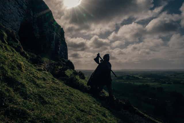 Un caballero con capa y un hacha al hombro desciende a contraluz por la ladera de una montaña.