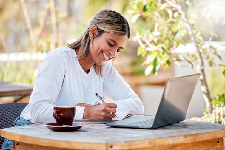 A young woman smiling and writing in a notebook while sitting at a cafe outdoors