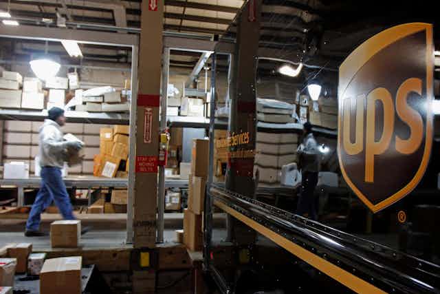 A worker carries boxes on  a loading dock toward a UPS truck.
