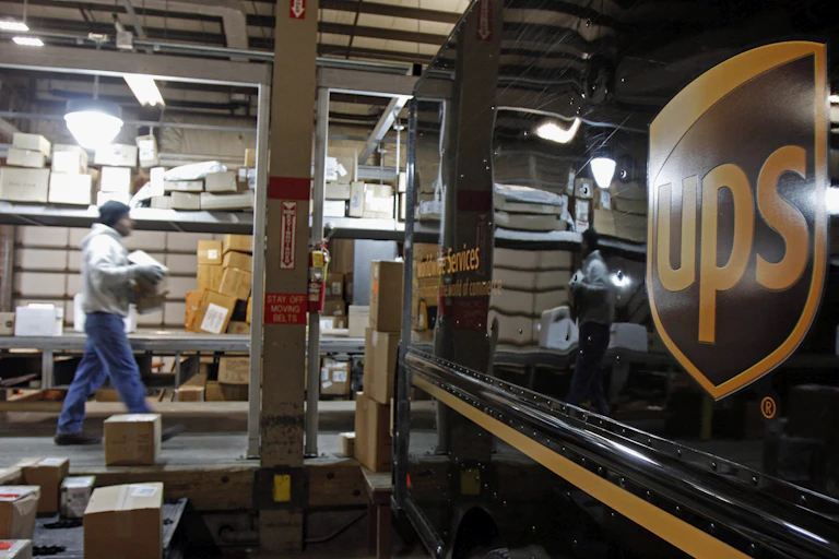 A worker carries boxes on a loading dock toward a UPS truck.