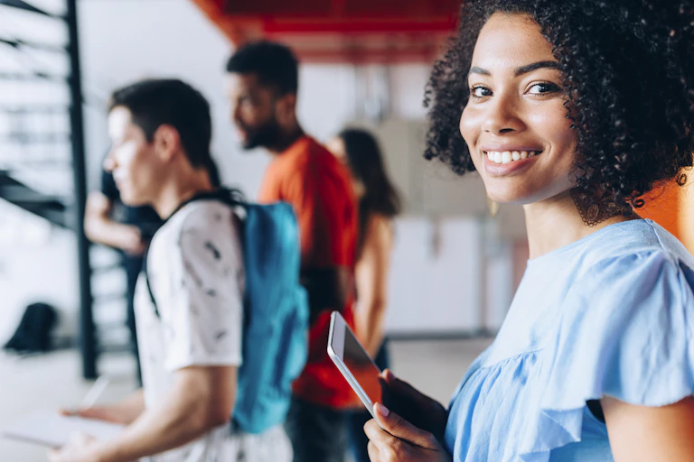 A Black female college student looks straight on as other college students move about in the background.